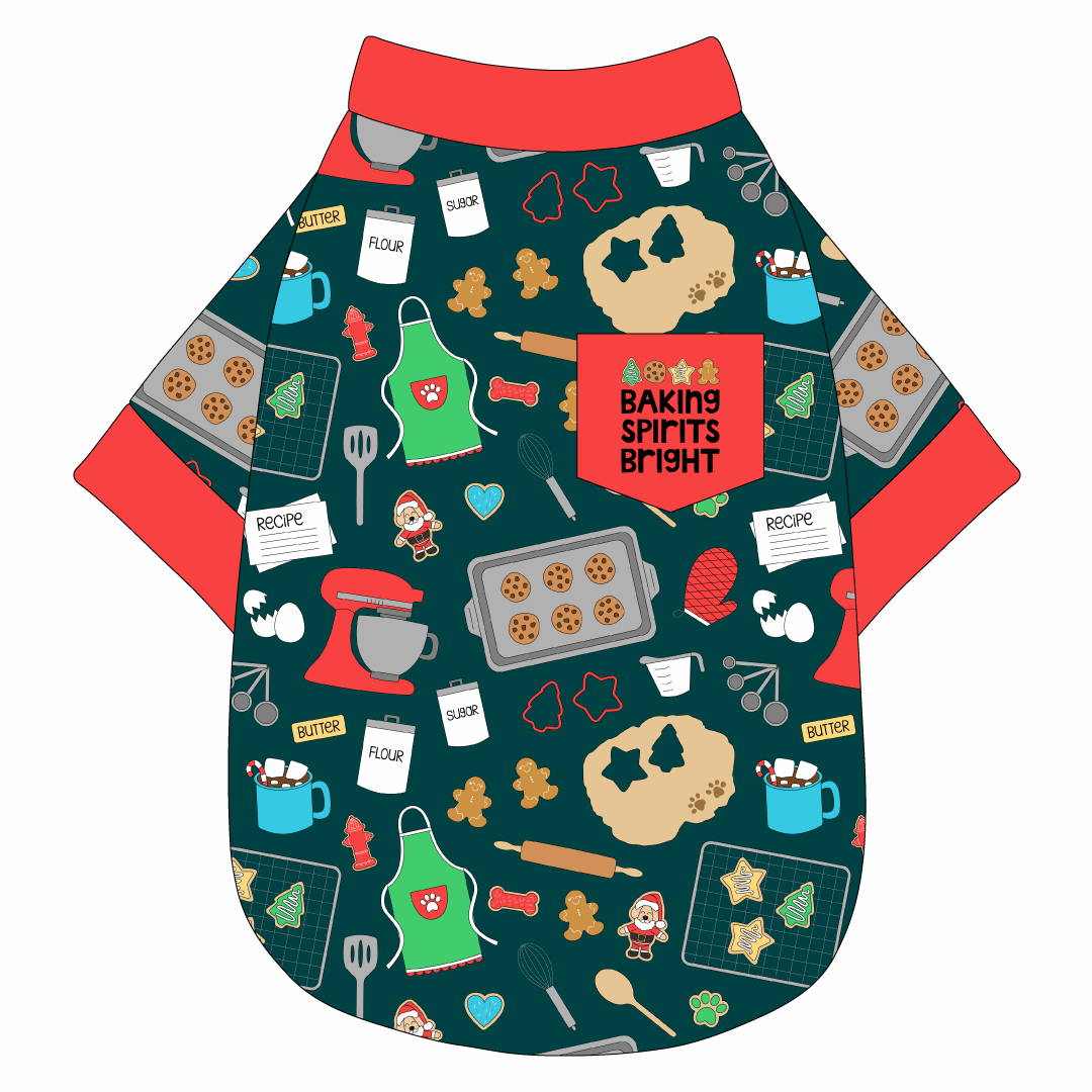 Tee shirt in a dog friendly pattern that shows cookies baking, a light green apron, recipe cards and more on a deep emerald background. It's complete with red trim on the neck, waist and sleeves, as well as a red pocket that reads Baking Spirits Bright.