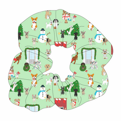 Scrunchie hair accessory in dog friendly pattern that shows the different dog breeds being naughty at Christmas.  Some are getting in gifts, some knocked over the tree, some are tangled in lights, and more. Design is on a light green background.